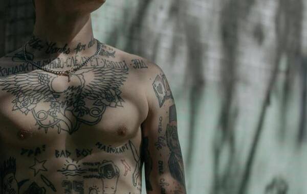Tattooed individual showcasing intricate body art, including script and symbolic designs, reflecting diverse tattoo culture. Soft lighting highlights the details and textures of the tattoos.