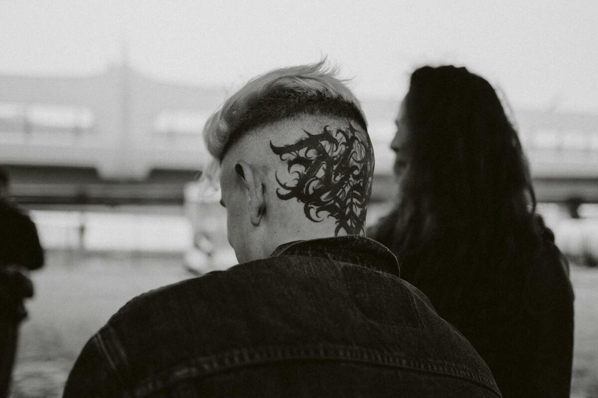 Close-up of a person with a detailed tattoo on the back of their head, showcasing intricate designs, embodying modern tattoo culture. A blurred figure is visible in the background, adding to the urban vibe of the scene.