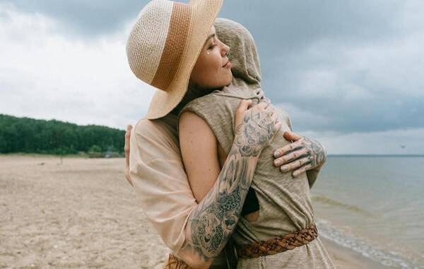 Two tattooed individuals embrace on a beach under a cloudy sky, highlighting the connection and expression of tattoo culture. One wears a wide-brimmed hat, while the other is partially concealed by a hooded garment.