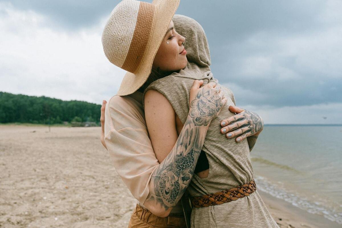 Couple embracing on a beach, showcasing intricate tattoo designs on their arms, with a cloudy sky and natural scenery in the background, reflecting the connection between tattoo culture and self-expression.