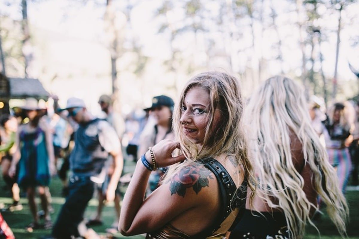 A woman with a colorful tattoo smiles at a lively outdoor festival, surrounded by dancing crowds and trees, showcasing vibrant tattoo culture and festival vibes.