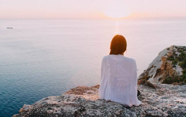 A solo female traveler enjoys a serene sunset view over the ocean while sitting on a rocky ledge, embodying the spirit of adventure and introspection