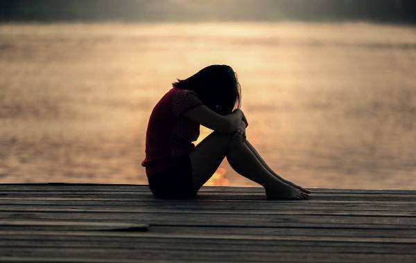 A silhouette of a solo female traveler sitting on a wooden dock, embracing her knees with a serene lake and sunset in the background, evoking a sense of reflection and solitude.