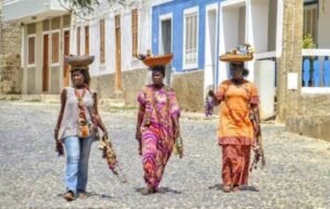 Three women carrying baskets on their heads walk down a cobblestone street in Cabo Verde, showcasing vibrant traditional attire and local floral garlands.
