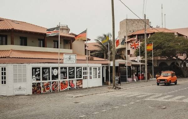 Street view of Cape Verde featuring colorful flags from various countries, a charming restaurant with a vibrant storefront, and an orange vintage car parked nearby. The setting captures the essence of local culture in a picturesque coastal town.