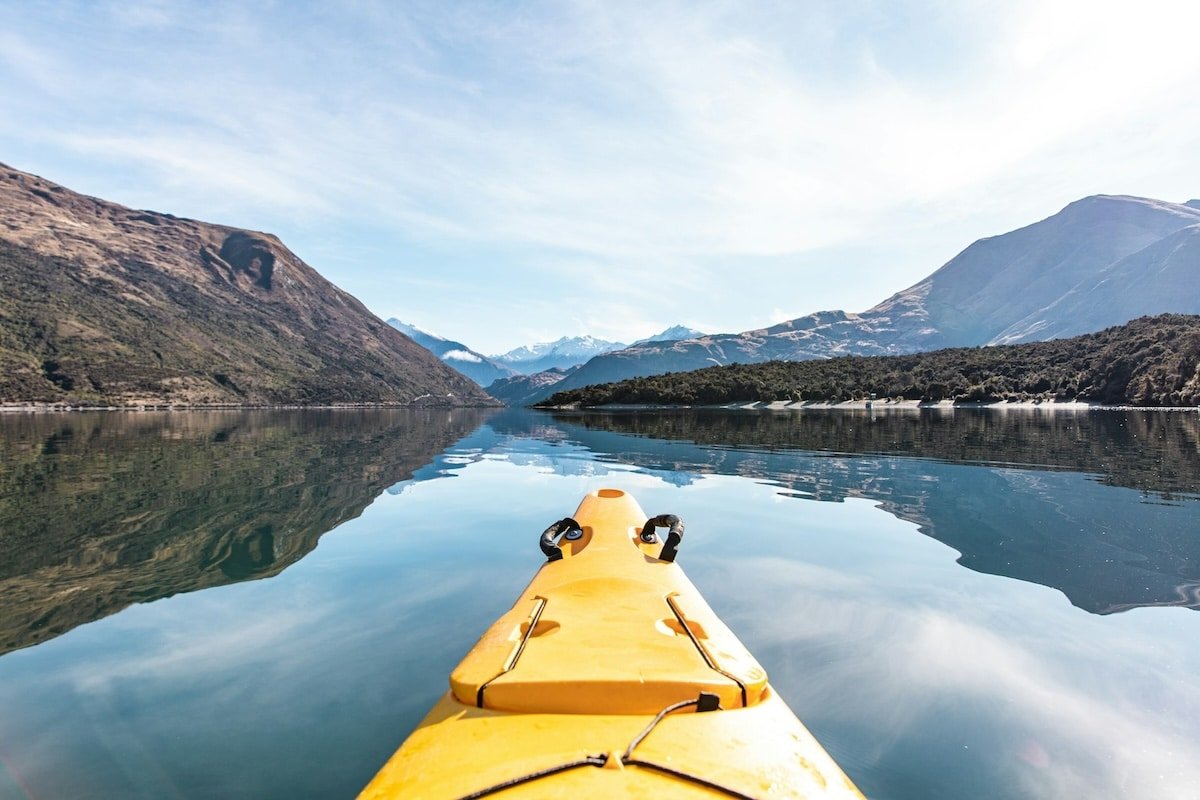 Kayaking through the calm waters of New Zealand's stunning landscape, featuring mountains and clear blue skies, with a focus on the yellow kayak in the foreground.