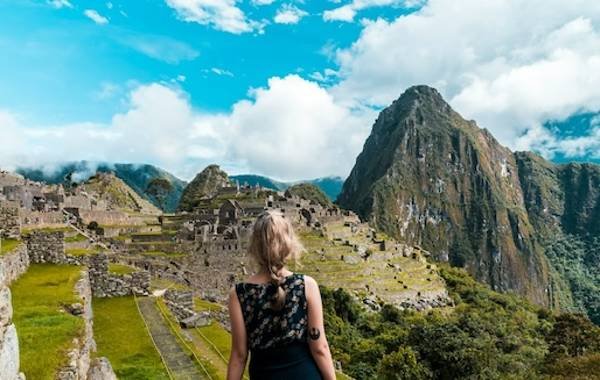 A woman gazes at the ancient ruins of Machu Picchu, surrounded by lush greenery and towering mountains under a bright blue sky with fluffy clouds.