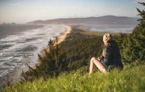 A solo traveler sits on a grassy hillside overlooking a scenic coastal view, featuring a winding beach and lush greenery under a clear sky.