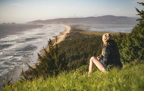 A solo traveler sits on a grassy hillside overlooking a scenic coastal view, featuring a winding beach and lush greenery under a clear sky.