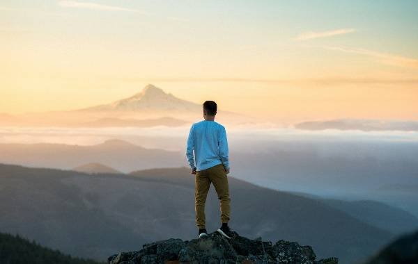 A solo traveler stands atop a rocky outcrop, gazing at a majestic mountain peak in the distance during a beautiful sunset. Layers of misty hills create a serene backdrop.