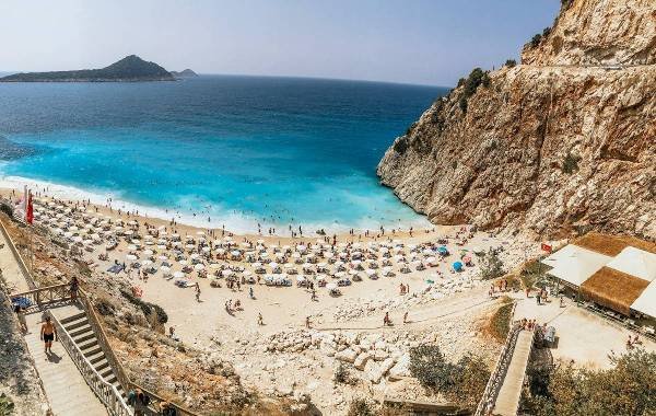 A scenic view of Antalya, Turkey, showcasing a beautiful beach with umbrellas and sunbathers, framed by a stunning turquoise sea and rocky cliffs in the background.