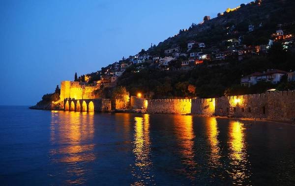 Night view of Antalya, Turkey, featuring illuminated historic walls reflecting on the water, with hillside homes visible in the background.