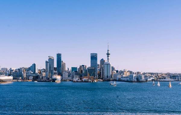 Skyline of Auckland, New Zealand, featuring modern skyscrapers and the iconic Sky Tower, with sailboats on the turquoise waters and a clear blue sky.