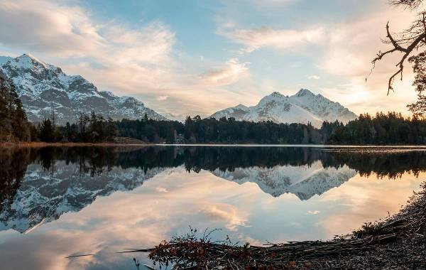 Stunning landscape of Bariloche, Argentina, featuring snow-capped mountains reflecting in a tranquil lake at sunset, surrounded by lush forests.