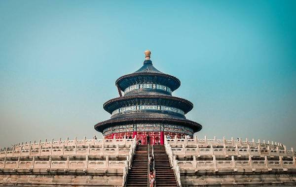 Temple of Heaven in Beijing, showcasing its iconic circular architecture and vibrant colors against a clear blue sky. The structure is surrounded by traditional stone railings and steps leading up to its entrance.