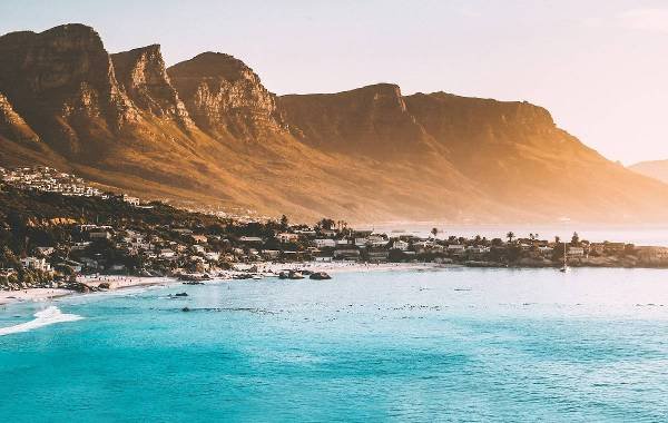 Scenic view of Cape Town, South Africa, featuring golden mountains, a tranquil beach, and clear turquoise waters at sunset.