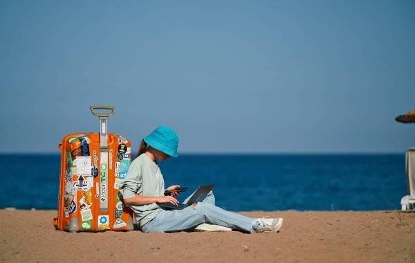 Person working on a laptop by the beach with an orange suitcase.