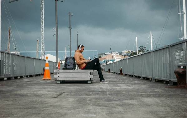 Person sitting on a bench on a dock working on a laptop with a backpack and headphones.