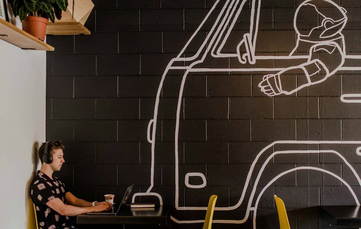 Person wearing headphones working on a laptop with a coffee cup in a modern cafe featuring a mural of an astronaut.
