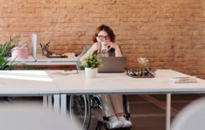 Person in wheelchair working on laptop in modern, open office setup with exposed brick wall and greenery