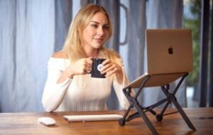 Woman in white long-sleeve shirt working on a laptop at a wooden desk while holding a coffee mug, representing digital nomad lifestyle.