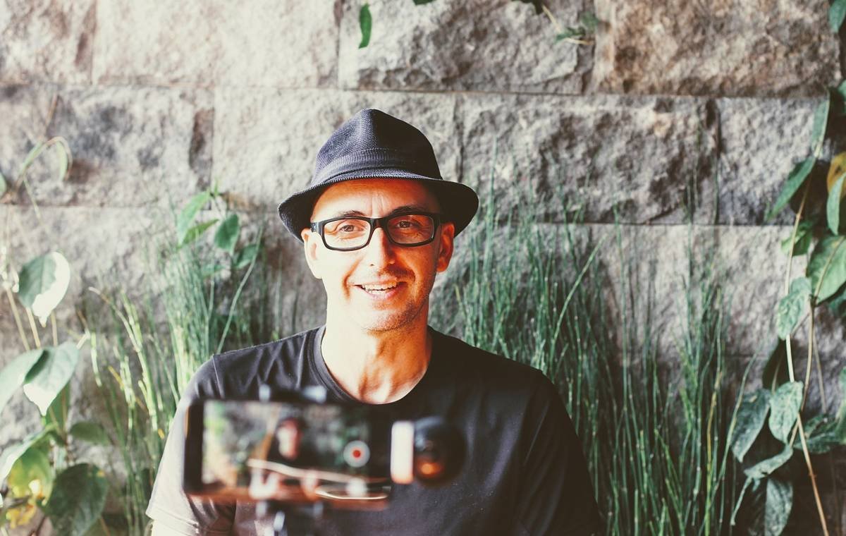 Man wearing a hat and glasses recording a video for a vlog in an outdoor setting with a stone wall and greenery in the background