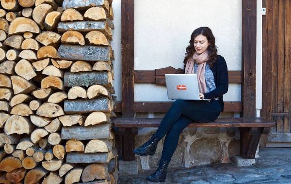 Woman working on a laptop outdoors beside a stack of firewood