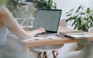 Person working remotely on a laptop at a desk with plants and notebooks in the background