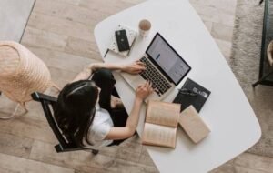 Person working remotely on laptop at a bright, minimal desk with books, smartphone, and coffee mug.