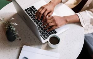 Person typing on a laptop at a round table with a coffee cup and tablet, representing digital nomad lifestyle