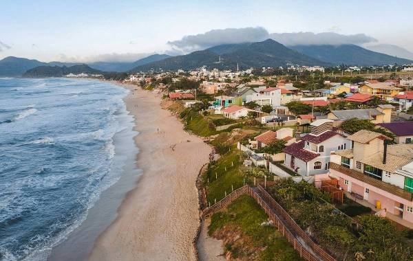 Aerial view of a scenic beach in Florianópolis, Brazil, showcasing golden sands, gentle waves, and residential areas nestled against lush green hills and mountains in the background.