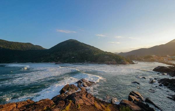 Breathtaking coastal view of Florianópolis, Brazil, showcasing rolling waves, rocky shorelines, and lush green mountains under a vibrant sky. Ideal for nature lovers exploring Brazilian landscapes.