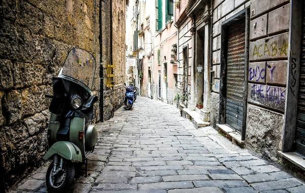 Narrow cobblestone street in Genoa, Italy with parked scooters and historic buildings.