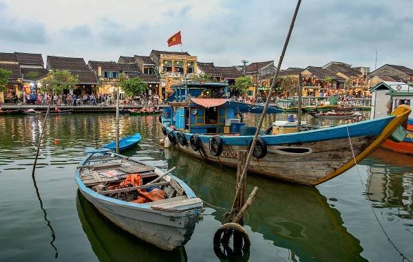 Colorful boats docked on the river in Hoi An, Vietnam, with a vibrant streetscape and Vietnamese flag in the background, capturing the charm of this historic town.