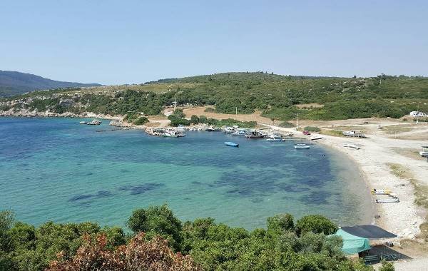 Scenic view of a tranquil beach in Izmir, Turkey, featuring clear blue waters, boats anchored near the shore, and lush greenery surrounding the sandy coastline.