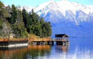 Scenic view of Neuquén, Argentina, showcasing a peaceful lake with a wooden pier surrounded by towering mountains and lush trees, reflecting the stunning landscape in the clear water.