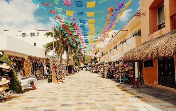 Colorful papel picado decorations hang above a vibrant market street in Playa del Carmen, Mexico, lined with shops selling local crafts and attire, under clear blue skies.