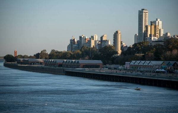 City skyline of Rosario, Argentina, featuring modern high-rise buildings alongside the Paraná River, with historic warehouses and people enjoying the waterfront promenade.