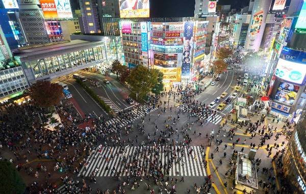 Birdeye view of Tokyo traffic with many billboards, flashing lights and pedestrians walking chaotically around the square