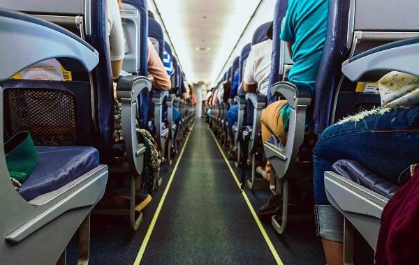 View of a central aisle in an airplane with passengers seated on both sides, featuring rows of blue seats and overhead storage compartments.