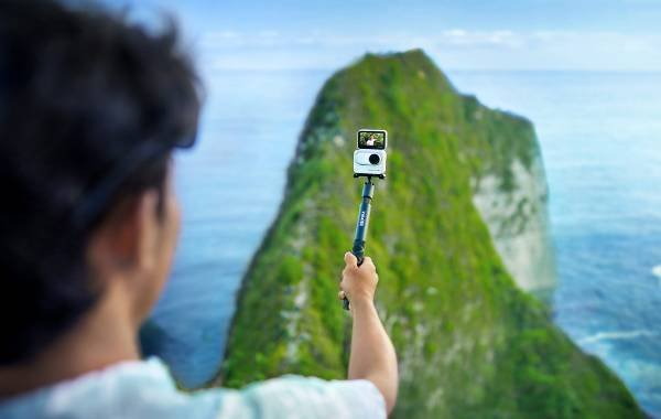 Person using an action camera on a selfie stick to capture scenic view of a green cliff and ocean background with advanced Insta360 gadget