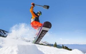 Snowboarder performing a jump while holding an Insta360 action camera during a sunny day on a snowy mountain slope.