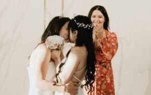 A newlywed LGBT couple shares a kiss at their wedding ceremony, with a woman applauding in the background.