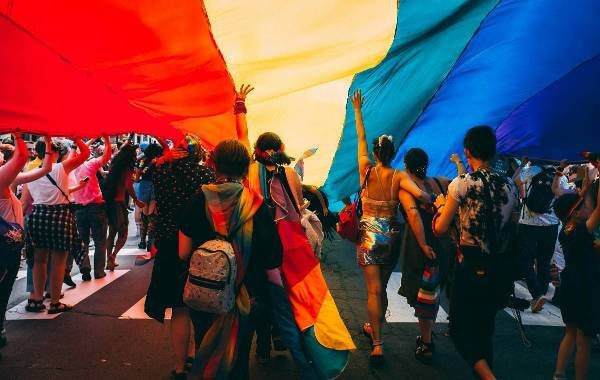 LGBT pride parade with diverse crowd celebrating under large rainbow flag.