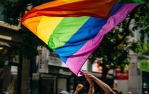 People raising a large, vibrant rainbow flag during an LGBT pride parade in a city setting.