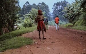 A child in a red hoodie and wraps stands on a dirt road in Gitega, Burundi, holding a bag, while another person walks in the background among lush greenery.