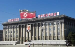 Government building in Pyongyang, North Korea, featuring a large North Korean flag and a prominent portrait of a leader, with people walking in front on a clear day.