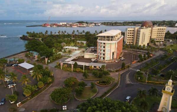 Aerial view of Apia, Samoa, featuring vibrant greenery, modern buildings, and the picturesque coastline with boats in the harbor under a partly cloudy sky.