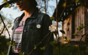Woman in a denim jacket with a daisy pin stands beside flowering branches in a serene outdoor setting in Romania, with a rustic wooden building in the background