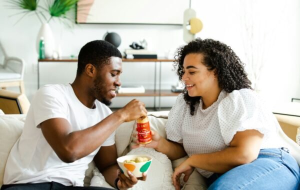 A couple enjoying takeout food together in a cozy living room, sharing a snack and a drink while smiling at each other, highlighting the joy of food delivery and shared meals